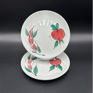 Könnte beinhalten: Zwei weiße Keramikteller mit handgemalten Designs. Jeder Teller zeigt ein rotes Obst- und grünes Blattdesign. Die Teller sind übereinander gestapelt, mit einem silbernen Ständer dazwischen.