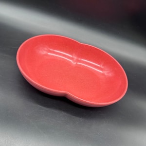 May include: A red, kidney-shaped serving dish. The dish has a smooth, slightly textured surface and a rounded rim. The color is a vibrant, solid red. The dish is empty and sits on a dark surface.