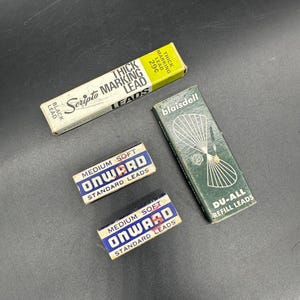 Może przedstawiać: Cztery pudełka z zapasowymi ołówkami do ołówków mechanicznych. Pudełka są w różnych kolorach i mają różne marki. Pudełka są oznaczone jako "Scripto Thick Marking Lead", "Onward Medium Soft Standard Leads", "Onward Medium Soft Standard Leads" i "Blaisdell Du-All Refill Leads".