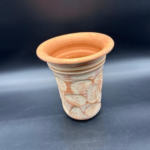 May include: A handmade terracotta vase with a flared rim and a textured exterior. The vase features a repeating seashell pattern carved into the clay. The rim and interior are a warm orange-brown color, while the exterior has a lighter, weathered appearance.