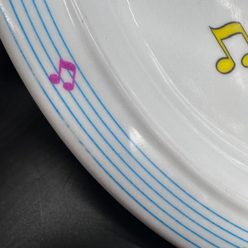 以下が含まれることがあります： 青い平行線と音符が描かれた白いお皿のクローズアップ。音符はピンクと黄色です。お皿は縁がカーブしており、光沢のある仕上がりです。