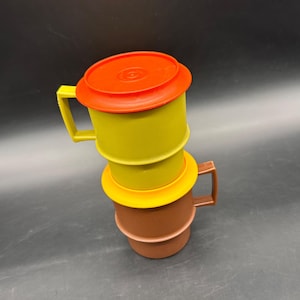 May include: Two stacked plastic mugs with handles. The top mug is olive green with a bright orange lid. The bottom mug is brown with a yellow rim. Both mugs feature a cylindrical shape with a distinct horizontal ridge around the center body.