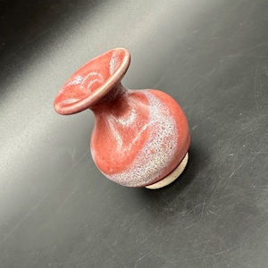 Puede incluir: Un pequeño jarrón de cerámica roja con un borde acampanado y un cuerpo redondeado. El jarrón tiene un patrón moteado blanco y una base de color crema. El jarrón mide aproximadamente 10 cm de alto.