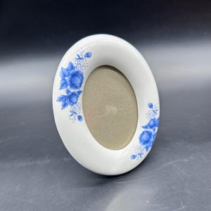 Puede incluir: Marco de fotos ovalado de cerámica blanca con diseños florales azules. El marco tiene un interior de color neutro y está decorado con flores y hojas azules en la parte superior e inferior.