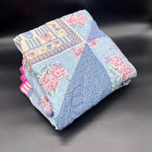 May include: Folded floral quilt with a patchwork design. The quilt features a light blue background with pink roses and other floral patterns. The edges are trimmed with a pink and white striped fabric.