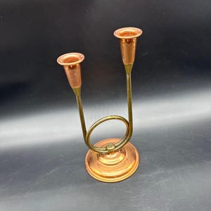 May include: A copper-colored candle holder with two candle cups. The candle holder has a circular base and a curved, brass-colored stem that supports the candle cups. The candle cups are a lighter copper color than the base.