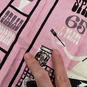 Pu&ograve; includere: Tessuto rosa con un motivo a griglia nera e illustrazioni a tema cibo. Il tessuto presenta la parola "BREAD" e il numero "65". Sono visibili anche una forchetta e un barattolo.