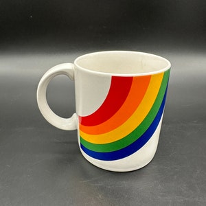 May include: White ceramic mug with a rainbow design. The rainbow is curved and has red, orange, yellow, green, blue, and purple stripes.