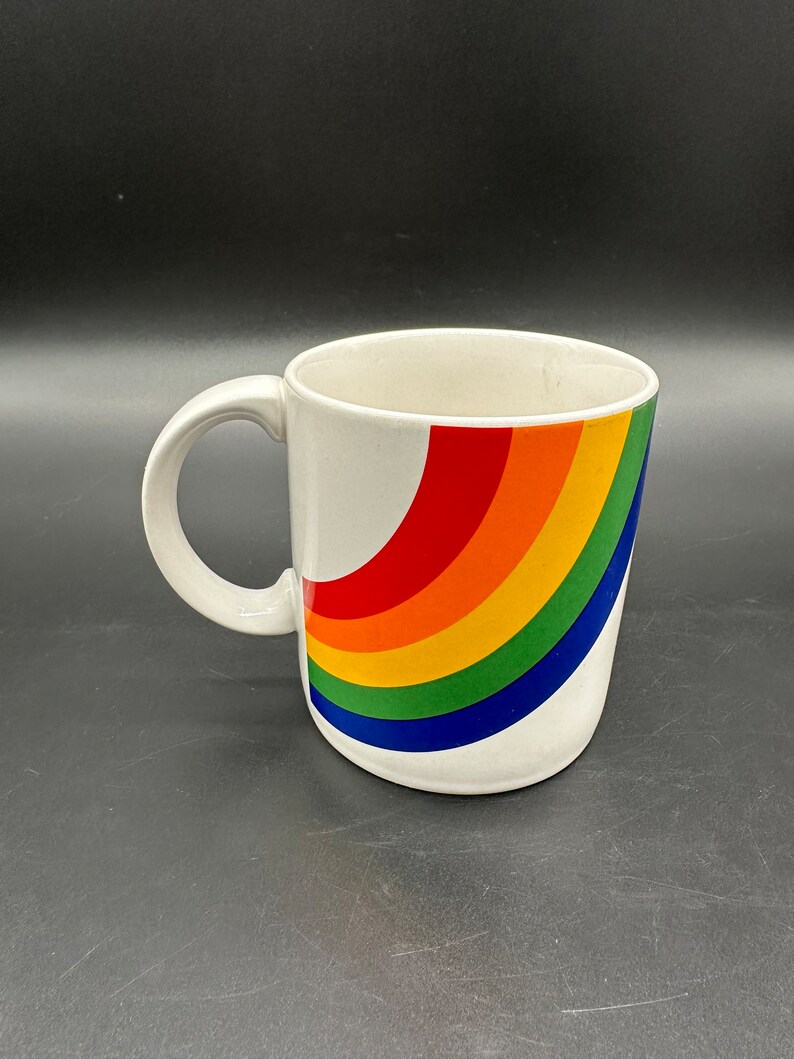 May include: White ceramic mug with a rainbow design. The rainbow is curved and has red, orange, yellow, green, blue, and purple stripes.