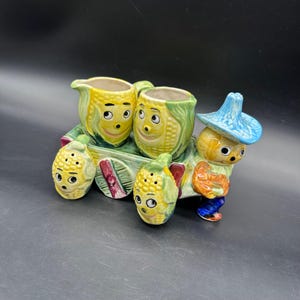 May include: A whimsical ceramic corn-themed salt and pepper set with a matching creamer and sugar bowl. The set includes two corn-shaped salt and pepper shakers, a creamer, and a sugar bowl, all on a small cart with a scarecrow figure.