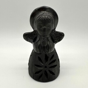 May include: A dark brown, carved figurine of a girl wearing a hat and dress. The dress features a large, stylized flower design. The girl's arms are outstretched, and her eyes are closed. The figurine is on a white background.