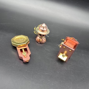 Może przedstawiać: Trzy miniaturowe metalowe przedmioty w stylu steampunk. Obiekty obejmują zegar, latarnię i prostokątne pudełko z mosiężnymi i miedzianymi odcieniami. Zegar ma ośmiokątną tarczę. Latarnia ma kopulasty wierzch. Pudełko ma element cylindryczny.