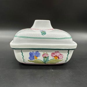 May include: A rectangular white ceramic butter dish with a matching lid. The exterior features hand-painted floral motifs in blue, pink, and green, with green wavy vertical lines and a horizontal green band encircling the base and lid rim.