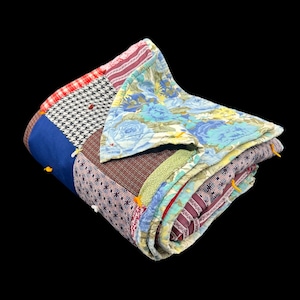 Puede incluir: Un edredón de retazos plegado con una variedad de patrones y colores de tela. El edredón presenta secciones de telas florales azules, pata de gallo y a rayas. Se ven detalles de hilo amarillo.