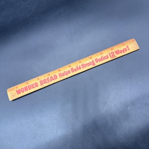 May include: A vintage wooden ruler with red text that reads "WONDER BREAD Helps Build Strong Bodies 12 Ways!" The ruler is marked with inch measurements, and is approximately 30 cm long.