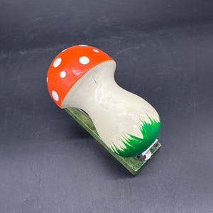May include: A whimsical mushroom-shaped stapler. The stapler features a red cap with white polka dots, a cream-colored stem, and a green base. The stapler is a unique desk accessory.