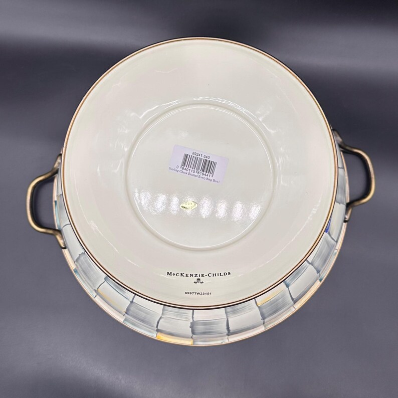 Op de afbeelding: Een ronde emaille kom met twee messingkleurige handgrepen aan de zijkanten. De buitenkant heeft een grijs en wit ruitpatroon. De cr&egrave;mekleurige onderkant toont de tekst MacKenzie-Childs, een barcode-sticker en een klein rond goudkleurig label.