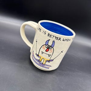 May include: White ceramic mug with a blue rim. The mug has a cartoon monster skiing with the text "Life is better when you ski" on the side.