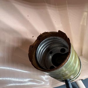 K&ouml;nnte beinhalten: Nahaufnahme einer Vintage-Lampenfassung mit Metallinnenraum und abgenutztem, braunem &Auml;u&szlig;eren. Die Fassung ist an einem schwarzen Draht befestigt und vor einem hellrosa Hintergrund platziert.