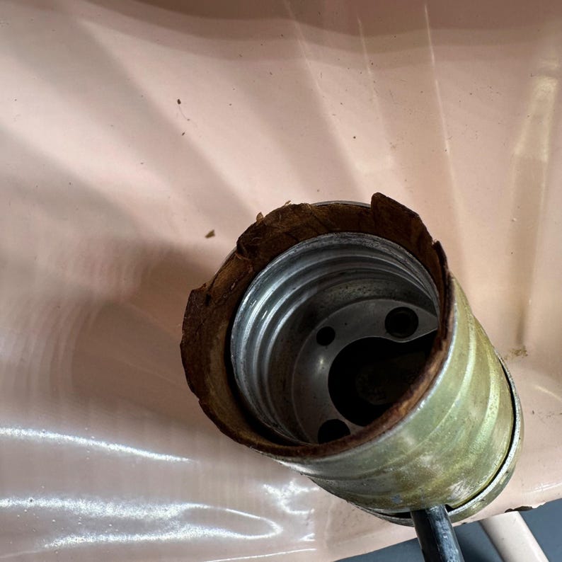 K&ouml;nnte beinhalten: Nahaufnahme einer Vintage-Lampenfassung mit Metallinnenraum und abgenutztem, braunem &Auml;u&szlig;eren. Die Fassung ist an einem schwarzen Draht befestigt und vor einem hellrosa Hintergrund platziert.