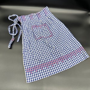 May include: A blue and white gingham apron with red embroidered details. The apron has a large pocket and ties at the waist.