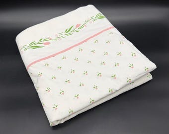 May include: A folded white bed sheet featuring a delicate pattern of small pink flowers and green leaves. A horizontal pink stripe separates the main section from a border decorated with a larger floral vine motif in pink and green.