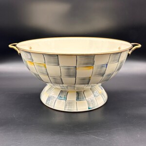 Op de afbeelding: Een kom op voet met een cr&egrave;mekleurige binnenkant en een buitenkant met een handgeschilderd ruitpatroon in grijze, blauwe en gele tinten. De kom heeft een metalen goudkleurige rand en twee bijpassende gebogen handvatten aan weerszijden.