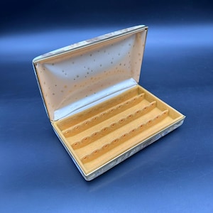 May include: Open jewelry box with a gold and cream patterned exterior. The interior is lined with a peach-colored fabric and has multiple slots for storing jewelry. The box is rectangular and has a hinged lid.