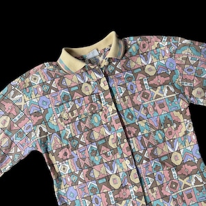 May include: A button-down shirt with a geometric pattern in pastel colors. The shirt features a light yellow collar with striped accents and two chest pockets. The pattern includes shapes in pink, blue, brown, and yellow.