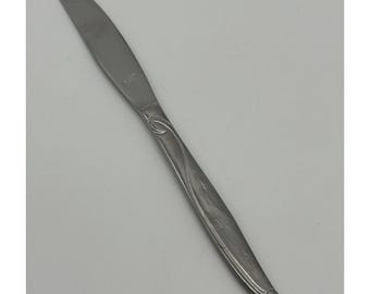 May include: A stainless steel butter knife with a decorative handle. The knife has a smooth, curved blade and a handle with a stylized floral design. The handle also features small starburst patterns.
