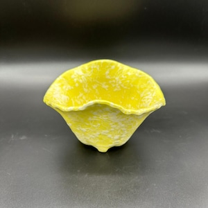 May include: A yellow ceramic bowl with a ruffled edge and a speckled white pattern. The bowl has a flared shape and three small feet. The background is black.