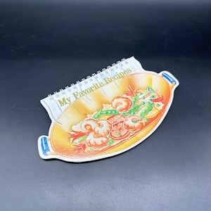 Puede incluir: Un cuaderno espiralado con una cubierta blanca y una ilustración colorida de un wok lleno de camarones, verduras y arroz. La cubierta dice "Mis recetas favoritas".