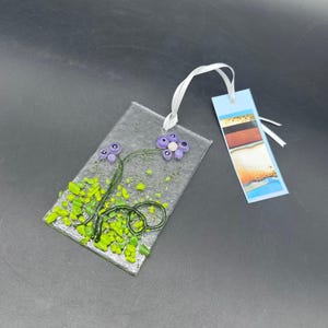 May include: A clear glass bookmark with a floral design. The bookmark features purple flowers, green leaves, and a white ribbon. A separate blue bookmark with abstract art is attached.