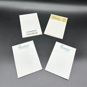 May include: Four white notepads with horizontal lines are displayed on a dark surface. Two pads have the text "New Prometrium PROGESTERONE USP" in green. One pad has "CombiPatch" and another has "Estradab" printed on them.
