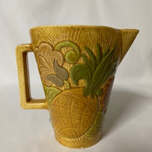 May include: A tan ceramic pitcher with a rectangular handle and a spout. The pitcher is decorated with embossed green, brown, and tan foliage and a pineapple design. The pitcher is approximately 8 inches tall.
