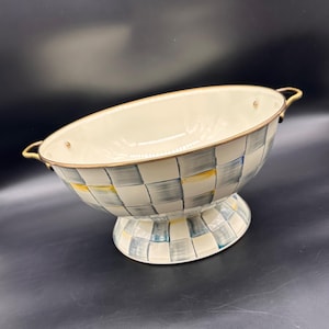 Op de afbeelding: Een ovale kom op een voet met een handgeschilderd ruitpatroon in blauw-, grijs- en geeltinten op een gebroken witte achtergrond. De kom heeft een metallic goudkleurige rand en twee bijpassende handgrepen aan de zijkant met een gladde afwerking.