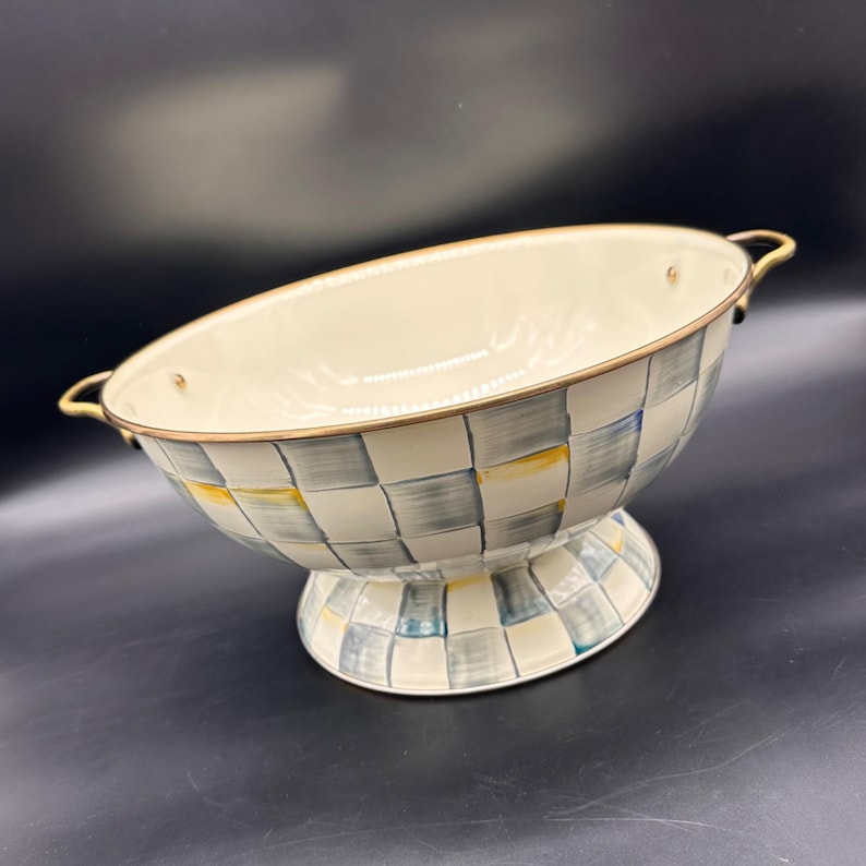 Op de afbeelding: Een ovale kom op een voet met een handgeschilderd ruitpatroon in blauw-, grijs- en geeltinten op een gebroken witte achtergrond. De kom heeft een metallic goudkleurige rand en twee bijpassende handgrepen aan de zijkant met een gladde afwerking.