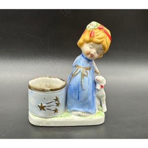 Puede incluir: Figura de porcelana vintage que representa a una niña con un vestido azul con un cordero y un pequeño recipiente. La niña tiene el pelo rubio y lleva una cinta roja. El recipiente es azul claro con detalles dorados y diseños de estrellas.