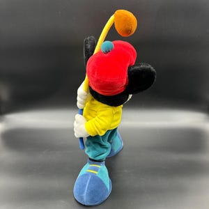 Puede incluir: Un juguete de peluche de Mickey Mouse con un sombrero rojo y una camisa amarilla, sosteniendo un palo de golf con una bola marr&oacute;n en el extremo. El juguete est&aacute; de pie sobre una superficie azul.