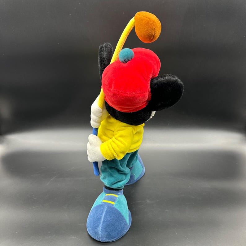 Puede incluir: Un juguete de peluche de Mickey Mouse con un sombrero rojo y una camisa amarilla, sosteniendo un palo de golf con una bola marr&oacute;n en el extremo. El juguete est&aacute; de pie sobre una superficie azul.