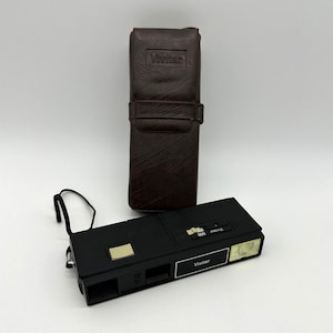May include: A black Vivitar 600 camera with a flash and a brown leather case. The camera has a rectangular shape with a silver accent and the Vivitar brand name. The case is tall and rectangular with a strap.