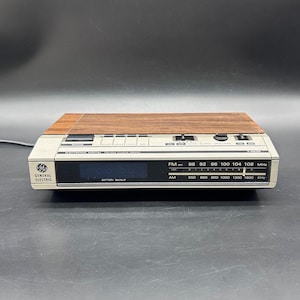 Puede incluir: Una radio despertador digital FM/AM General Electric vintage. La radio tiene una parte superior con vetas de madera y una pantalla digital. La cara de la radio muestra la hora y las frecuencias de radio. El texto "BATTERY BACKUP" es visible.