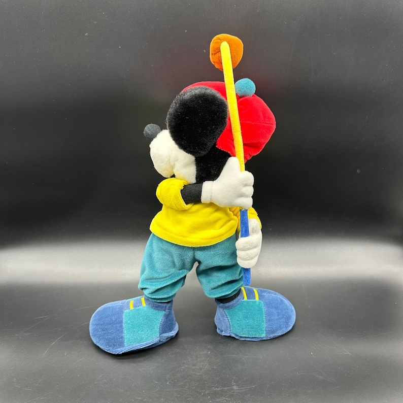 Puede incluir: Un mu&ntilde;eco de peluche de Mickey Mouse con una camisa amarilla, pantalones azules y un sombrero rojo. Est&aacute; sosteniendo un palo de golf con un eje amarillo y una bola roja en la parte superior.