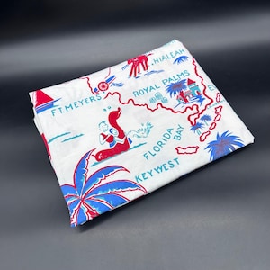 May include: White fabric with a red and blue map of Florida, featuring locations like Key West, Ft. Meyers, and Royal Palms. The map includes illustrations of palm trees, a cannon, and a mermaid-like figure. The fabric is folded.
