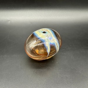 May include: A small, round, glass vase with a swirl pattern in shades of brown, blue, and white. The vase has a small opening at the top.