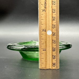 May include: A vintage green glass ashtray with a circular design. The ashtray is approximately 3 inches in diameter. The ashtray is made of a transparent green glass. The ashtray is on a dark surface.