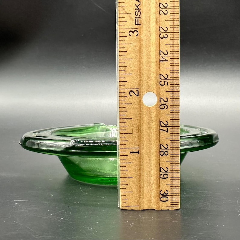 May include: A vintage green glass ashtray with a circular design. The ashtray is approximately 3 inches in diameter. The ashtray is made of a transparent green glass. The ashtray is on a dark surface.