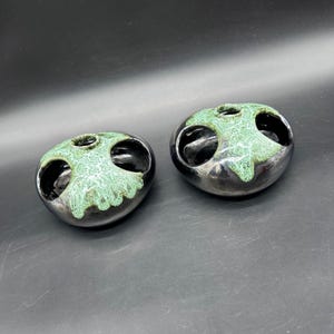 May include: Two ceramic ashtrays with a unique design. Each ashtray is a rounded shape with a glossy black finish and a textured, light green top. The ashtrays have three openings and a small hole in the center.