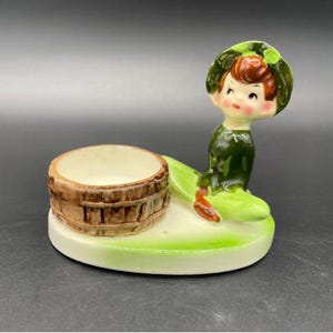 Puede incluir: Figura de cerámica vintage que representa una figura sentada con un vestido y sombrero verdes, sosteniendo un recipiente en forma de barril marrón. La figura está pintada en tonos verdes, marrones y crema, y se asienta sobre una base verde claro.