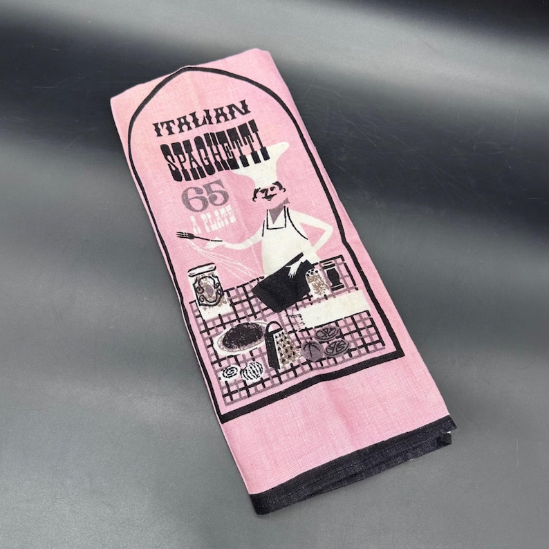 Pu&ograve; includere: Canovaccio da cucina rosa con bordo nero, con le parole "Italian Spaghetti" e un'illustrazione di uno chef. L'asciugamano ha anche i numeri "65" e le parole "A Plans".